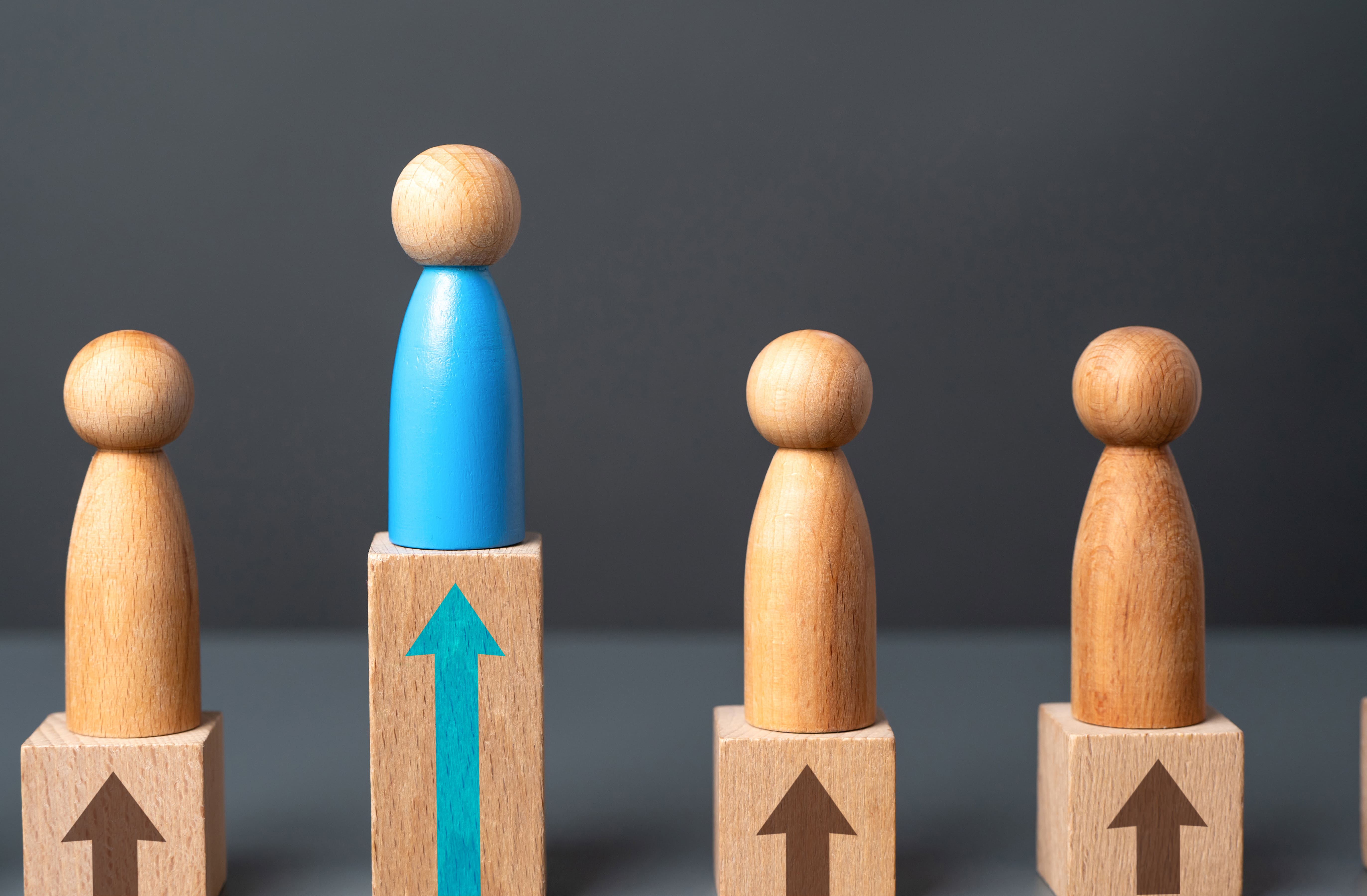 Four peg figures on pedestal blocks, one taller blue figure standing out from three uniform natural-wood figures, each block marked with an upward arrow, representing an operation that stands apart and scales above the rest